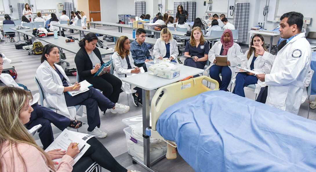 Learners in a hospital setting sitting in chairs around a preceptor in a white jacket who is standing.