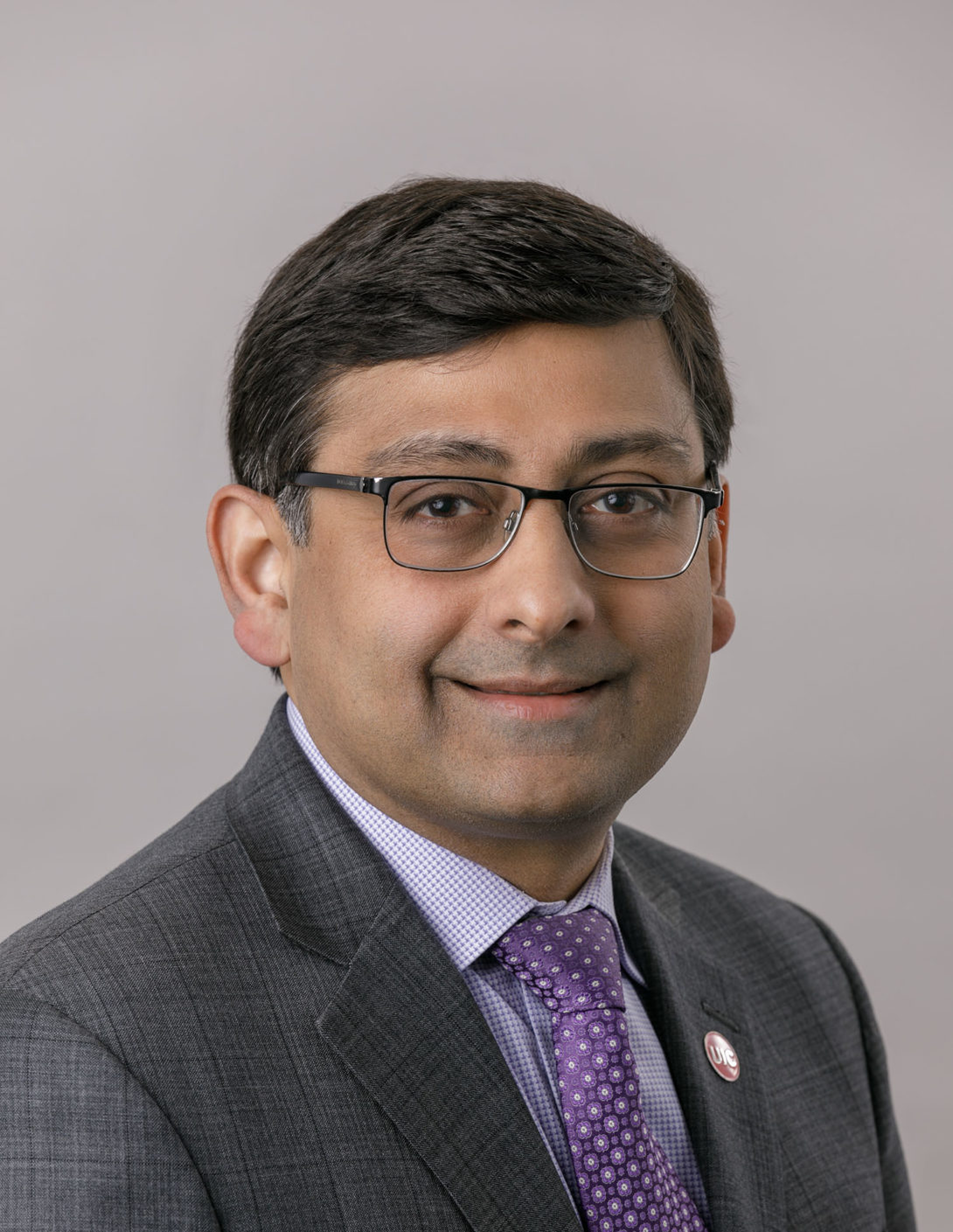 man with dark hair and glasses in a dark grey suit, light bliue shirt and lavender tie